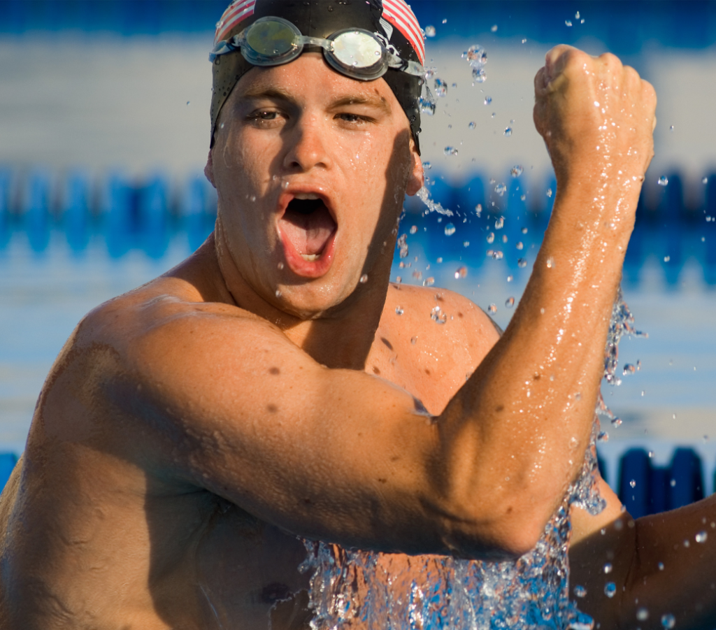 A swimmer wearing goggles and a swim cap celebrating with a raised fist and an open mouth while emerging from the water in a pool.